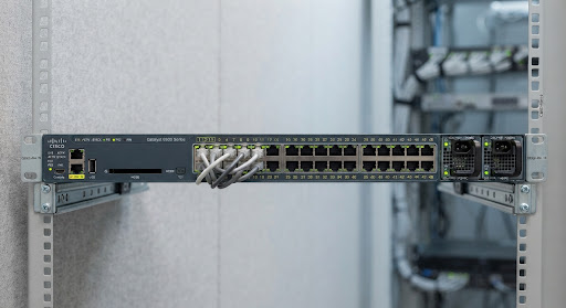 Front view photograph of Cisco Catalyst 9300 48-port enterprise network switch, showing RJ45 ports, LED status indicators, and modular power bay on a clean industrial neutral background with cool daylight illumination